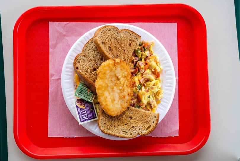 A breakfast platter with scrambled eggs, hash brown, toast, butter, and grape jelly on a red tray.