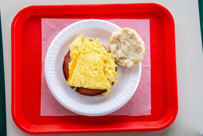 Breakfast sandwich with scrambled eggs, cheese, and sausage on a biscuit, on a white plate and red tray.